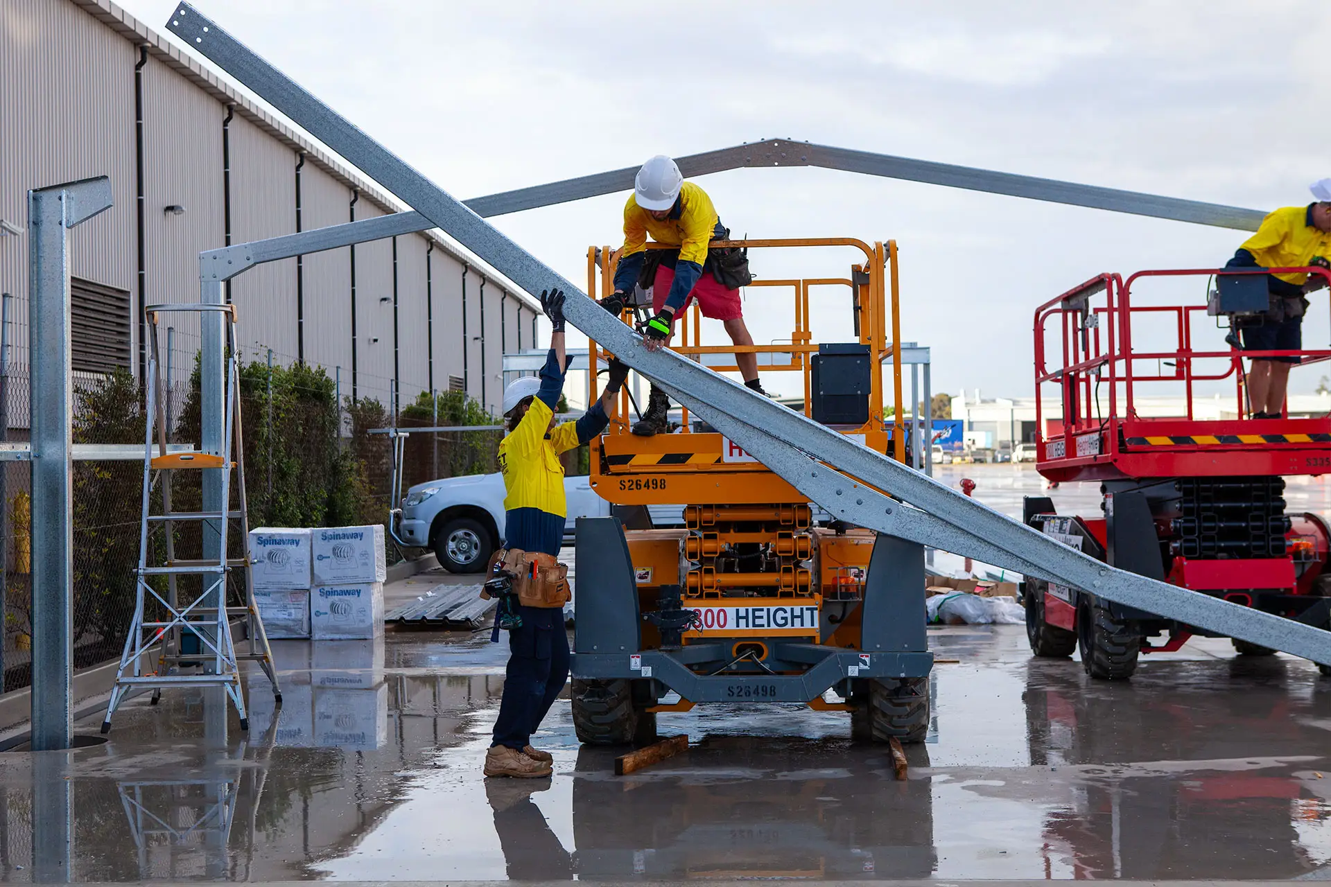 Two Debret workers lifting a large steel roofing piece during a commercial shed construction. One worker is elevated on a scissor lift, while another stretches from the ground to assist. A third worker on a separate scissor lift stands ready to receive the steel.