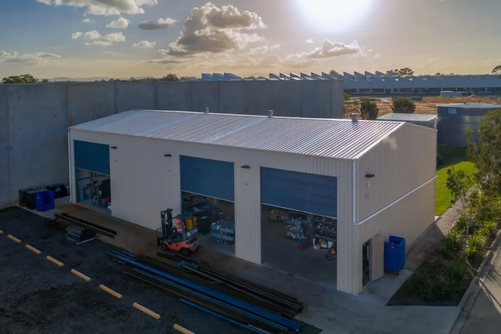 Wide drone shot of a purpose built commercial shed, showing skylights, whirlybirds, and three roller doors partially open with equipment inside. A forklift operator moves steel outside, while the PA door is open. The shed's COLORBOND® Paperbark® and Deep Ocean® finish stands out against the commercial landscape in the setting sun.