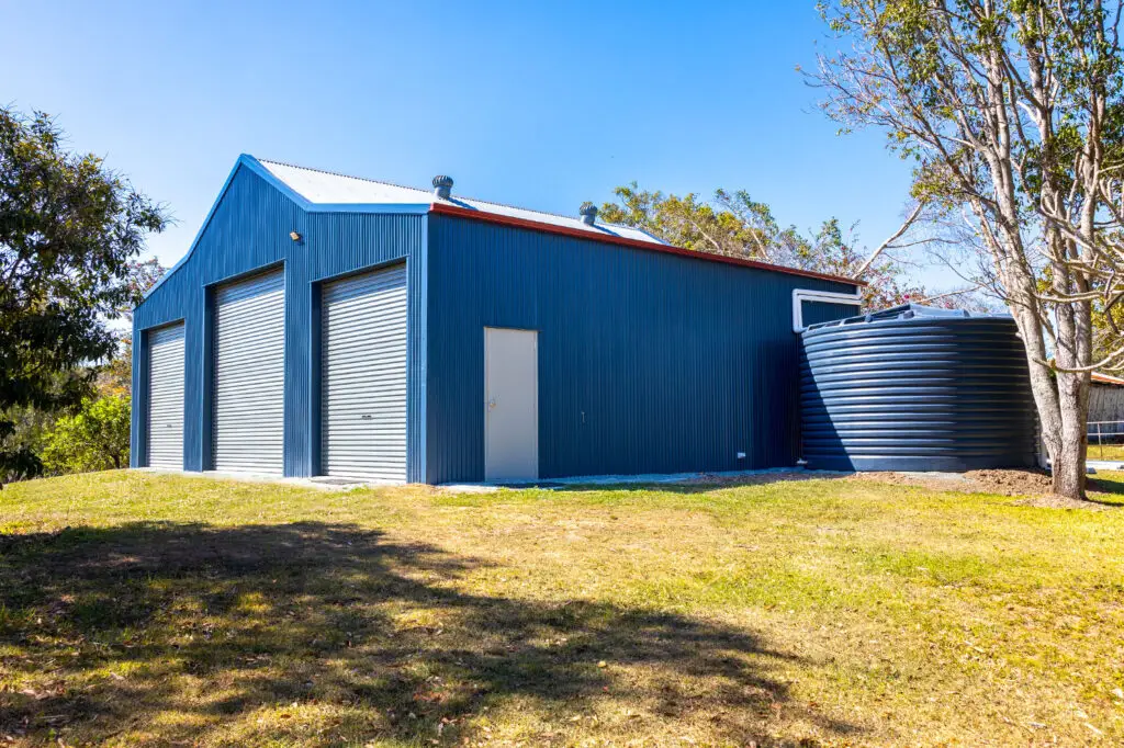 Side view of a custom Aussie barn in Draper, featuring COLORBOND® Deep Ocean® walls, Windspray® roller doors and roof, Manor Red® flashings, and a matching water tank with neat water management.