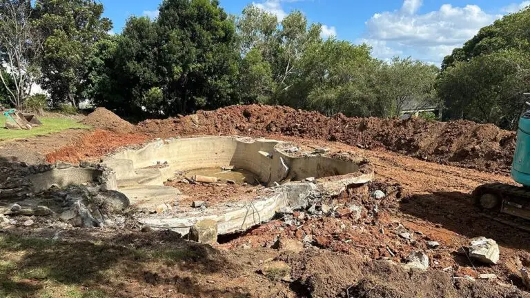Excavator breaking up old concrete pool during caravan and boat storage shed site preparation.