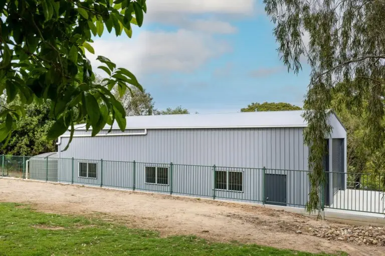 Side view of caravan and boat storage shed with green fence and mature trees in foreground.